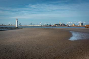 A lighthouse stands on a sandy beach under a blue sky, with an industrial city skyline in the background. The scene is calm and expansive.