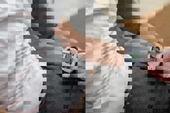 A person sits on a sofa wearing a white shirt, using a digital wrist blood pressure monitor displaying a reading. 