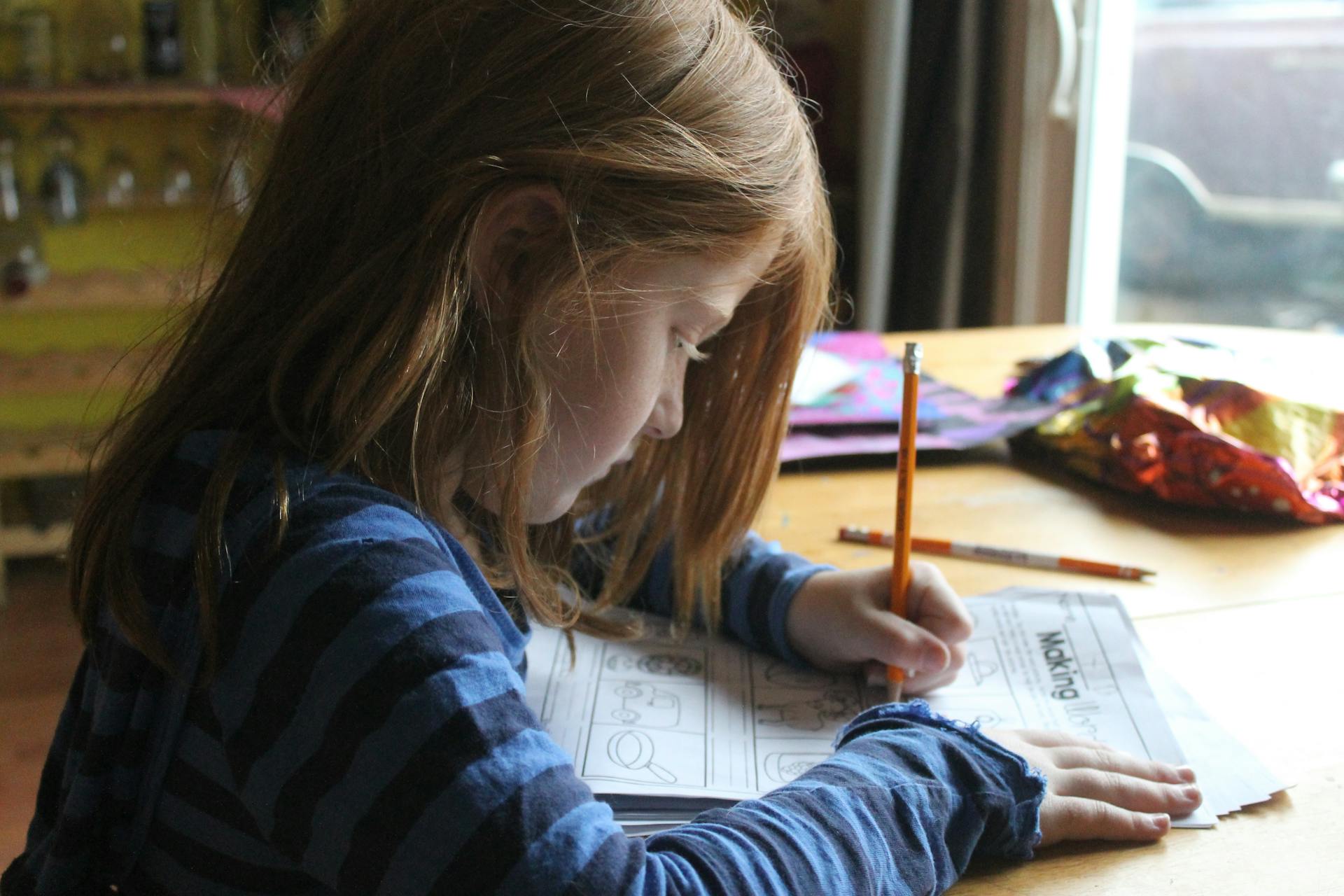 A young child with long hair is focused on doing homework at a wooden table. They're holding a pencil, colouring in a worksheet. 