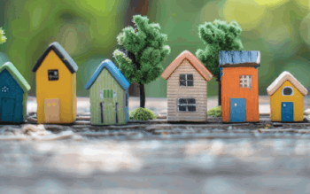 Miniature colourful houses with varied designs are lined up on a wooden surface, surrounded by small green trees.