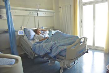 A person rests in a hospital bed, looking contemplative. Sunlight filters through large windows, creating a calm, serene atmosphere in the room.