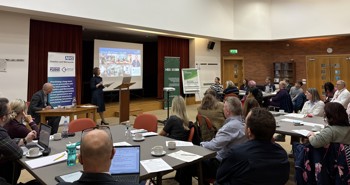 A speaker presents in a conference room to an engaged audience. Presentation slides and banners behind.