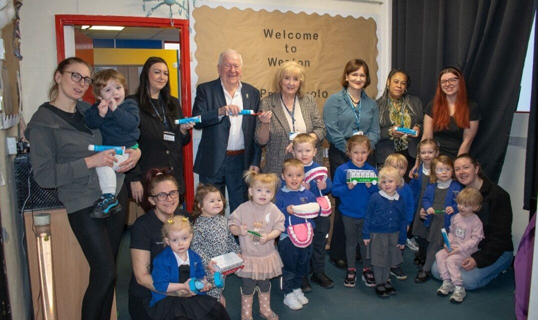 A group of smiling adults and children, holding dental care items, pose in a classroom. A sign on the wall reads, "Welcome to Weston." The scene conveys a cheerful and educational atmosphere.