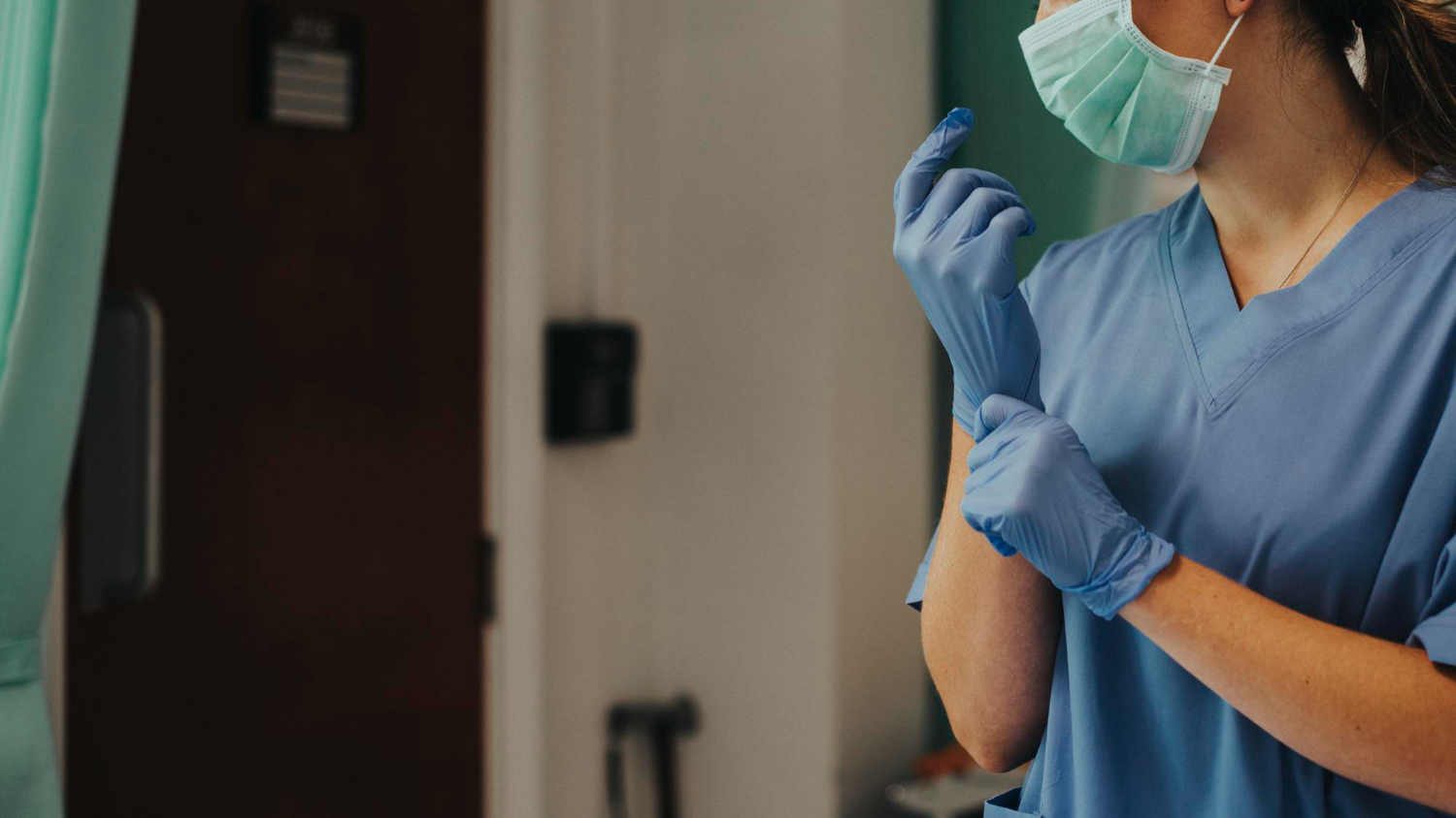 A healthcare worker in blue scrubs and a mask adjusts blue gloves, standing in a clinical setting. 