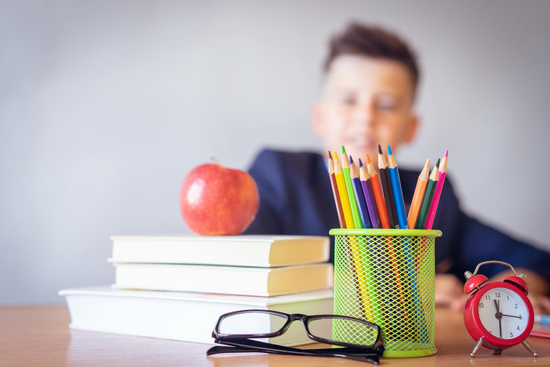 A child sits at a desk with colourful pencils in a green holder, an apple on stacked books, eyeglasses, and a red alarm clock.