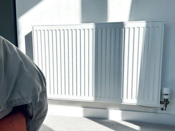 A white radiator casts shadows on a wall, with a corner of a beige fabric sofa partially visible on the left. Sunlight streams in.