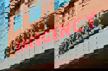 Red "EMERGENCY" sign on a hospital building facade with large windows and brickwork