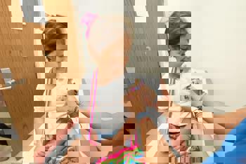 A young girl sits on a clinic bed, smiling at a nurse placing a colorful bandage on her arm. 