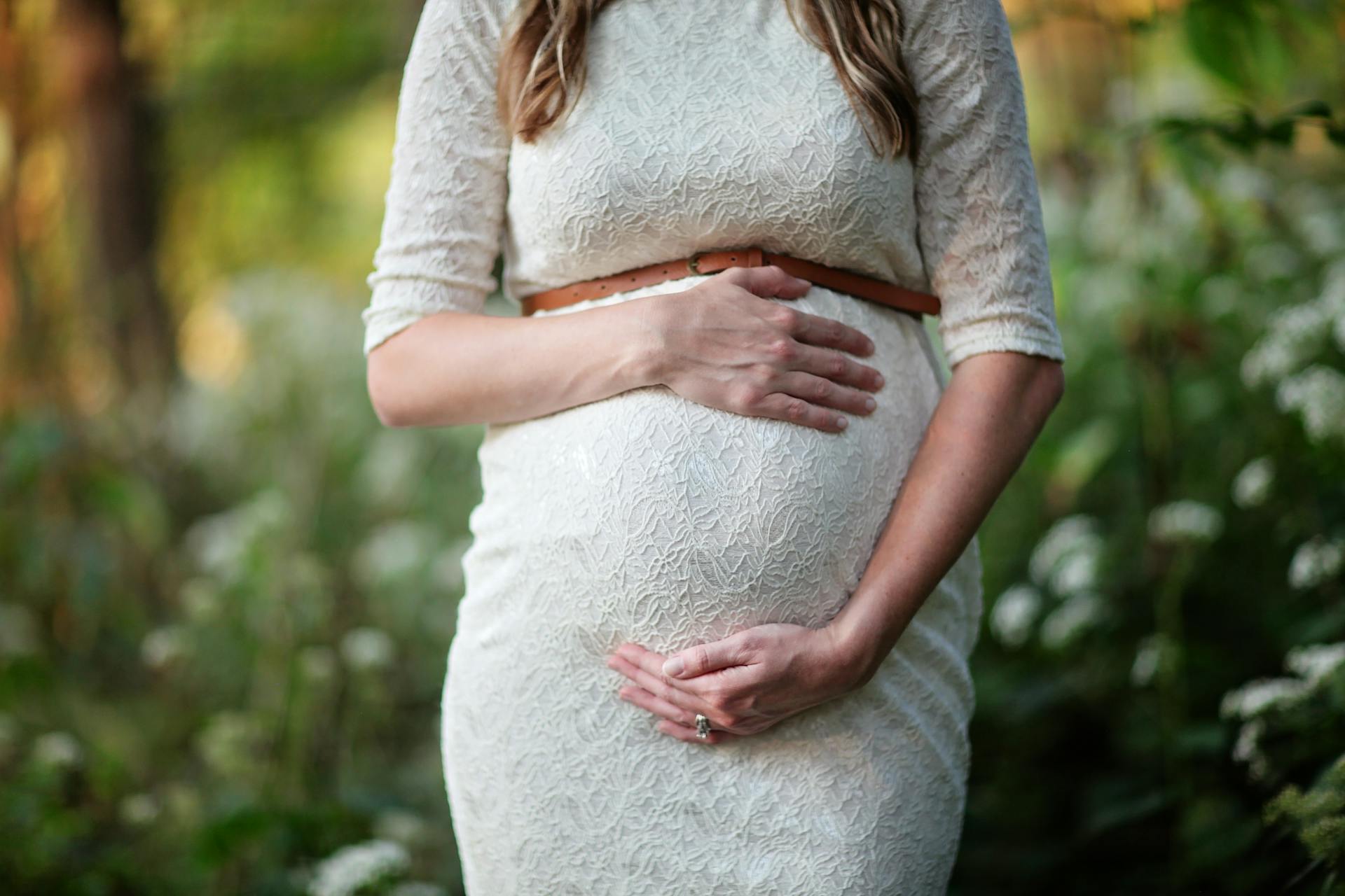 Pregnant woman in lace dress gently holds her belly with both hands, standing in a serene, green woodland setting.