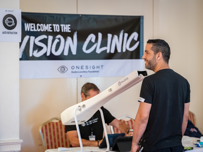 In the foreground, a man looking down a telescope-like box. In the background, a man sat at a desk with a banner above him reading "Welcome to the Vision Clinic"