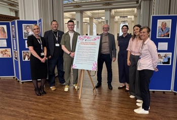 A group of seven people stands smiling beside an easel with text, at a photo exhibition titled "Living with Dementia." Blue display boards with portraits surround them in an elegant room. 