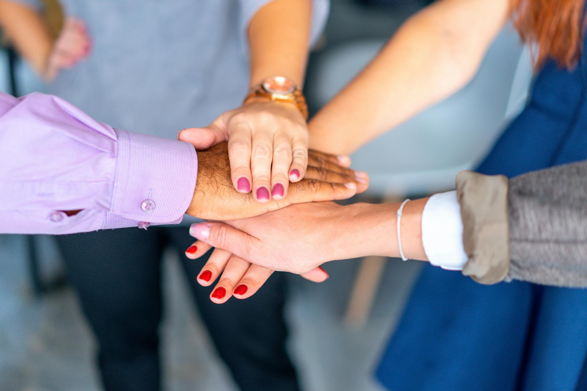 Five diverse hands with colorful nail polish are stacked together in a show of teamwork, conveying a sense of unity and collaboration.