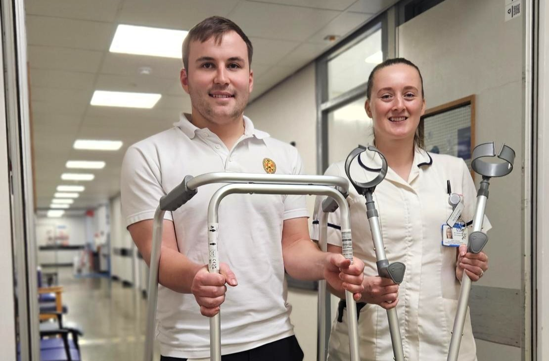 Two healthcare workers in uniform, smiling in a clinic hallway. One holds a walker, the other crutches. 