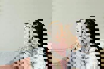 A woman gently kisses a baby on the cheek. The baby wears a pink dress and headband, while the woman is in a white shirt. 