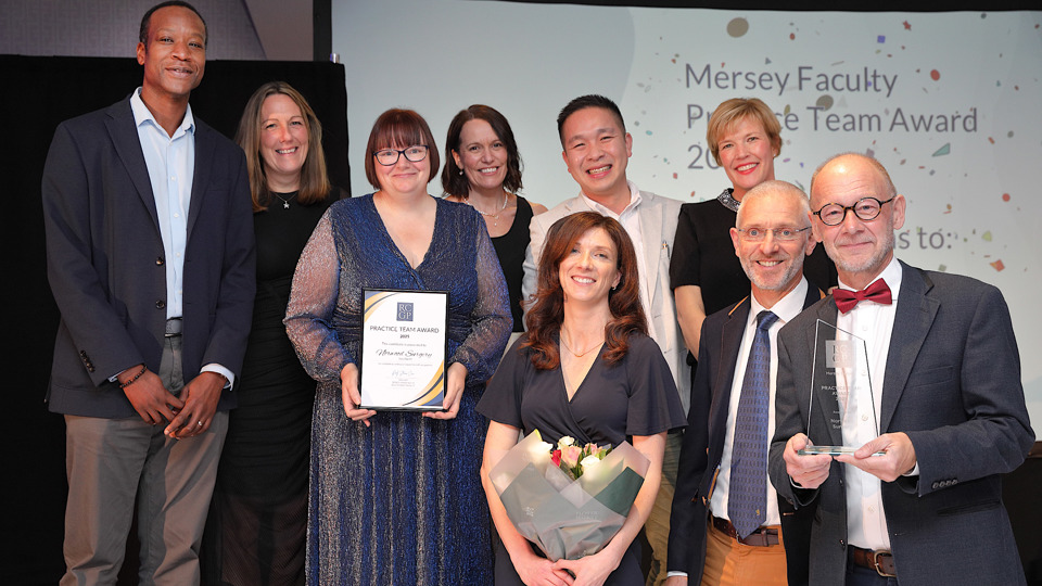A group of nine smiling people pose at an awards event. One holds a certificate and another holds a trophy. A projected display mentions "Mersey Faculty Practice Team Award 2025."